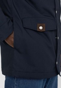 Navy jacket with buttoned pocket, featuring a brown leather accent on the pocket flap, made of sturdy fabric, with a smooth texture.