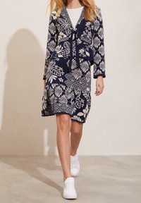 Námořnické šaty s květinovým vzorem a tříčtvrtečními rukávy, s vázáním vpředu a rozšířeným spodním okrajem, spárovány s bílými teniskami. Texturovaná látka.