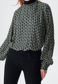 Blusa a maniche lunghe con colletto alto, motivo a onde nero e crema, con una vestibilità ampia e polsini arricciati, abbinata a jeans skinny neri.