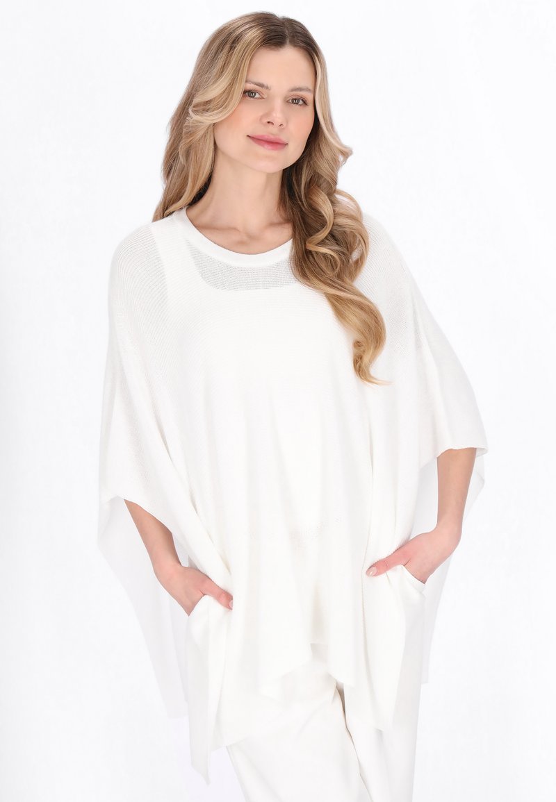 Donna con lunghi capelli biondi ondulati che indossa un poncho bianco lavorato a maglia morbido con tasche e pantaloni bianchi, in piedi davanti a uno sfondo bianco uniforme.