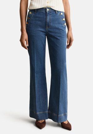 Hoch taillierte, weit geschnittene blaue Jeans mit goldfarbenen Knopfakzenten an den vorderen Taschen, getragen mit braunen spitzen Schuhen und einem in die Hose gesteckten weißen Oberteil.