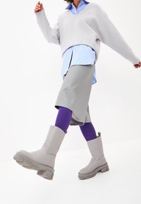 Suéter gris extragrande, camisa azul a rayas, falda skater gris, medias moradas y botines gris claro con suela gruesa. Fondo blanco.