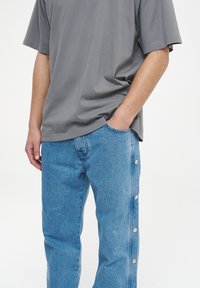 Camiseta gris de manga corta combinada con pantalones de mezclilla azul claro que cuentan con botones de presión metálicos a lo largo de la costura lateral exterior.