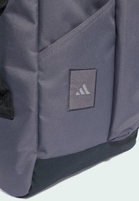 Šedý látkový batoh s texturovaným povrchem, jemným prošíváním a černým akcentním základem. Obsahuje výraznou nášivku s logem Adidas.