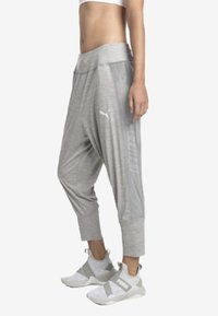 Vrouw in grijze Puma joggerbroeken en witte sportschoenen, staande tegen een effen witte achtergrond.