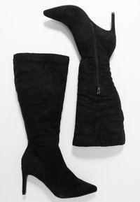 Par de botas negras de ante hasta la rodilla, con puntera afinada, cremallera lateral y tacones de aguja, dispuestas sobre un fondo blanco.