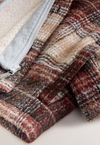 Capa exterior de forro polar con un patrón de cuadros en rojo, marrón y beige. Forro gris que complementa la cremallera y los detalles de costura. Textura suave.