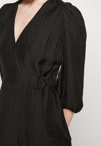Robe noire à envelopper avec texture à côtes verticales, encolure en V, manches bouffantes et une ceinture avec une boucle en métal ronde. Finition en tissu lisse.