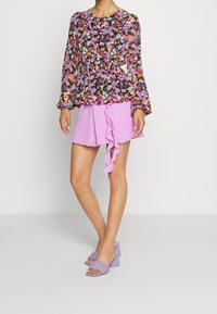 Blouse noire à motifs floraux à manches longues, associée à une mini-jupe à volants rose clair. Des sandales à talons blocs violettes complètent la tenue.