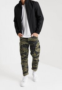 Svart zip-upp-jacka, vit T-shirt, kamouflage cargo-byxor med flera fickor, uppvikta muddar och vita sneakers.