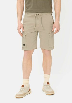 Mężczyzna w beżowych spodniach typu cargo z sznurkiem w pasie, w połączeniu z beżowo-brązowymi sneakersami i kremowymi skarpetkami na białym tle.