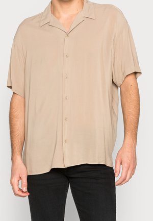 Mężczyzna w luźnej beżowej koszuli na krótki rękaw z guzikami i czarnych spodniach, stojący na tle jasnego, jednolitego tła.