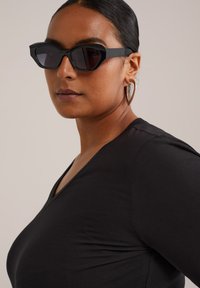 Una mujer de piel oscura y cabello recogido en un moño lleva unas gafas de sol negras con forma de ojo de gato, grandes pendientes en forma de corazón plateados y marrones, y una blusa negra con escote en V.