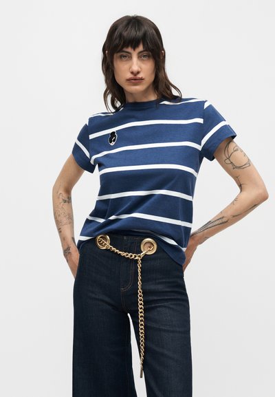 Γυναίκα με τατουάζ που φοράει μπλουζάκι με μπλε και άσπρες ρίγες και σκουρόχρωμο τζιν, διακοσμημένο με ζώνη αλυσίδα χρυσού, πόζα με τα χέρια στη μέση.