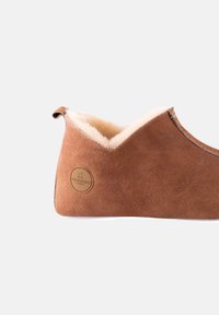 Pantufola in suede marrone con una morbida fodera in pelliccia bianca. Presenta una forma arrotondata e una piccola etichetta con il logo sul lato. Design minimalista.