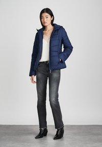 Moteris, vilkinti tamsiai mėlyną sintetinio audinio striukę per baltą blakstienų nėrinių viršų, su blukusiomis juodomis džinsinėmis kelnėmis ir juodais aulinukais, stovi ant pilko grindų.