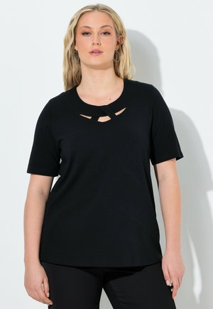 Donna bionda con orecchini a cerchio e top nero a maniche corte con dettaglio incrociato a taglio sullo scollo, in piedi davanti a un muro bianco.