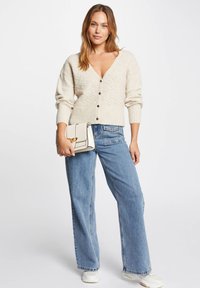 Beige cardigan med ett texturerat mönster och fem mörka knappar, parat med blå vida jeans och en krämfärgad handväska med guldhardware.