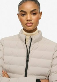 Lysbeige puffjakke med vandret syninger, høj krave og sort lynlåsdetalje. Modellen har en lys turtleneck på under.