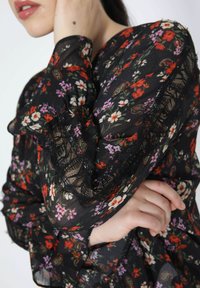 Black floral blouse with long sleeves. Features a mix of colorful floral patterns and lace trim. Lightweight, semi-sheer fabric with ruffle details.