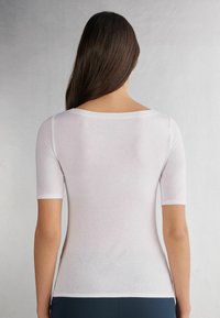 Hvid tætsiddende t-shirt med korte ærmer, en rund halsudskæring og glat tekstur. Tunike-længde der når til den høje hofte. Simpelt design, ingen mønstre.