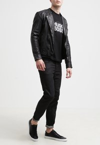 Chaqueta de motocicleta de cuero negro con textura acanalada, combinada con una camiseta negra que presenta un logo blanco, jeans negros ajustados y zapatillas negras.