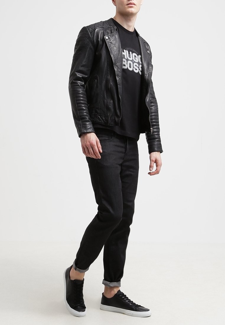 Chaqueta de motocicleta de cuero negro con textura acanalada, combinada con una camiseta negra que presenta un logo blanco, jeans negros ajustados y zapatillas negras.