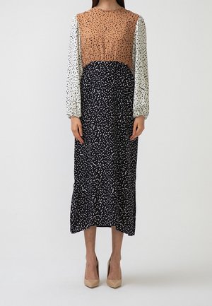 Mujer con un vestido de manga larga y bloques de color en tonos marrón, blanco y negro con patrones abstractos, lleva tacones altos beige y está de pie frente a un fondo liso.