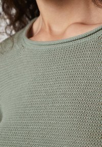 Gros plan d'une personne portant un pull en tricot vert clair, montrant le décolleté, la clavicule et des cheveux bouclés foncés.
