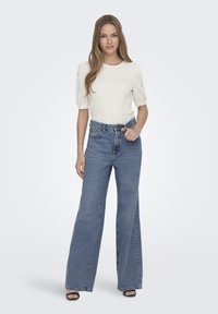 Top blanco texturizado con mangas abullonadas, combinado con jeans de mezclilla azul de tiro alto y pernera ancha. La modelo lleva sandalias de tacón negras.
