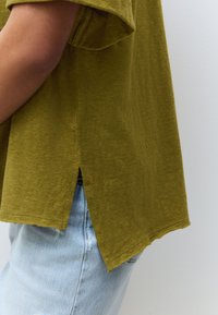 Olijfgroene T-shirt met korte mouwen en een losse pasvorm, voorzien van zijnaden met splitten en een zachte textuur, gedragen over lichtblauwe spijkerbroek.