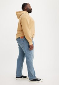 Beige hooded sweatshirt with a front pocket and light blue jeans. Black sneakers with white soles. Side view, neutral background.