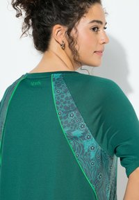 Zelené sportovní tričko s potiskem na zadní části s květinovým vzorem a kontrastním zeleným lemováním podél švů. Měkká, texturovaná tkanina.