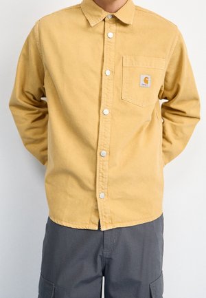 Mężczyzna noszący musztardowo-żółtą roboczą koszulę na guziki z przednią kieszenią i logo Carhartt, w połączeniu z ciemnoszarymi spodniami typu cargo.