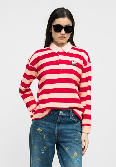 Γυναίκα που φοράει μαύρα γυαλιά ηλίου, ροζ και κόκκινη μπλούζα πόλο με οριζόντιες ρίγες και μικρό λογότυπο με πρόβατο, καθώς και μπλε τζιν με κεντήματα λουλουδιών.