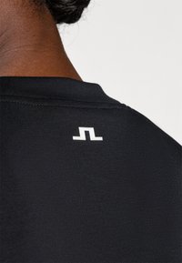 Fekete anyagú póló, fehér logóval a hátán, a nyakvonal közelében. Az anyag simának és testhezállónak tűnik.