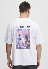 Hvid T-shirt med korte ærmer, der viser et grafisk print af palmer mod en pink- og lilla himmel, mærket "ARCHIVE" med fed sort tekst.
