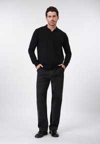 Μαύρο πόλο πουκάμισο με μακριά μανίκια και γιακά, συνδυασμένο με σκούρο γκρι τζιν παντελόνι ευθείας γραμμής και μαύρα παπούτσια, πόζα μπροστά σε απλό φόντο.