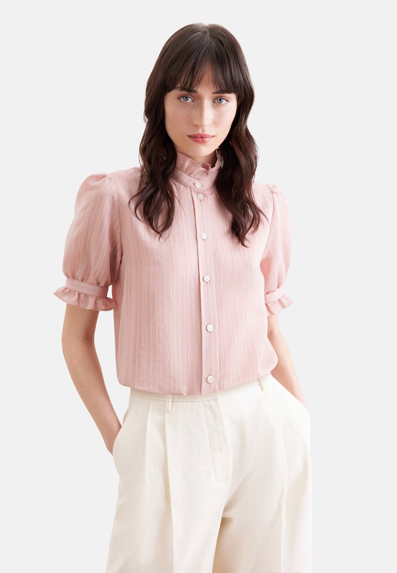 Jeune femme aux cheveux foncés portant un chemisier rose à manches courtes boutonné et un pantalon blanc taille haute, debout les mains dans les poches.