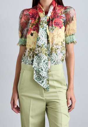 Femme portant un chemisier transparent floral à manches bouffantes et un foulard noué autour du cou, associé à un pantalon taille haute vert clair.
