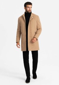 Cappotto tan con texture a righe verticali, tasche con zip e colletto alto, abbinato a un dolcevita nero e pantaloni neri slim-fit, e stivali neri.