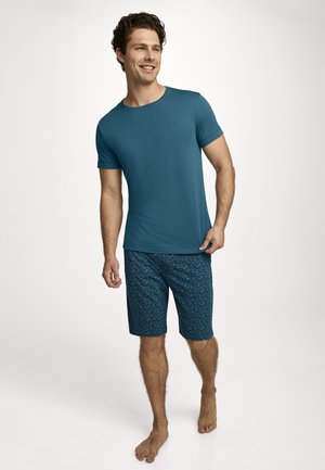 Smiling man walking barefoot, wearing a plain teal short-sleeve shirt and blue patterned knee-length shorts, on a white background.