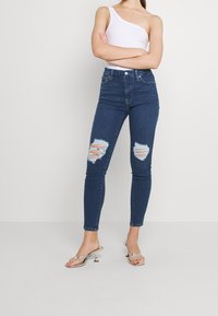 Jeans ajustados de color azul oscuro con detalles desgastados, que presentan grandes desgarrones en ambas rodillas, cintura alta y un diseño clásico de cinco bolsillos, combinados con sandalias plateadas.