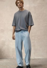 Młody mężczyzna z falującymi włosami, ubrany w luźny szary t-shirt, jasnoniebieskie spodnie o szerokich nogawkach i czarne buty na jednolitym beżowym tle.