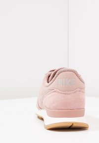 Růžové semišové tenisky s bílou mezipodešví a gumovou podrážkou. Mají polstrovaný límec a logo "NIKE" v světle šedé na patě.