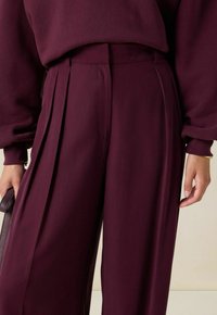 Vrouw in hoog-gewelfde, wijde, donkerpaarse broek en bijpassende lange mouw top, met een kleine handtas in haar linkerhand.