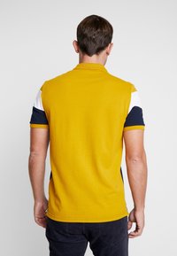 Homme debout de dos, portant un polo jaune avec des blocs de couleur blanc et bleu marine sur les manches et un pantalon foncé sur un fond uni.