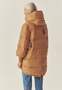 Brūna pūkaina jaka ar kapuci, ar diviem izšūtiem attēliem mugurā. Ribotas piedurknes un regulējams apakšmalas auklas, quilted dizains.