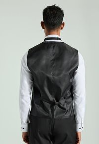 Zwarte vest met een gestructureerde afwerking, met een strik aan de achterkant, gedragen over een witte blouse met dubbele manchetten en zwarte knopen, gezien vanachter.