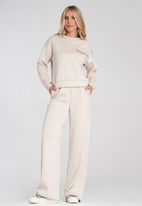Jasno beżowa bluza i dopasowane szerokie spodnie wykonane z miękkiego materiału. Bluza ma przednią kieszeń i ściągacze przy mankietach.